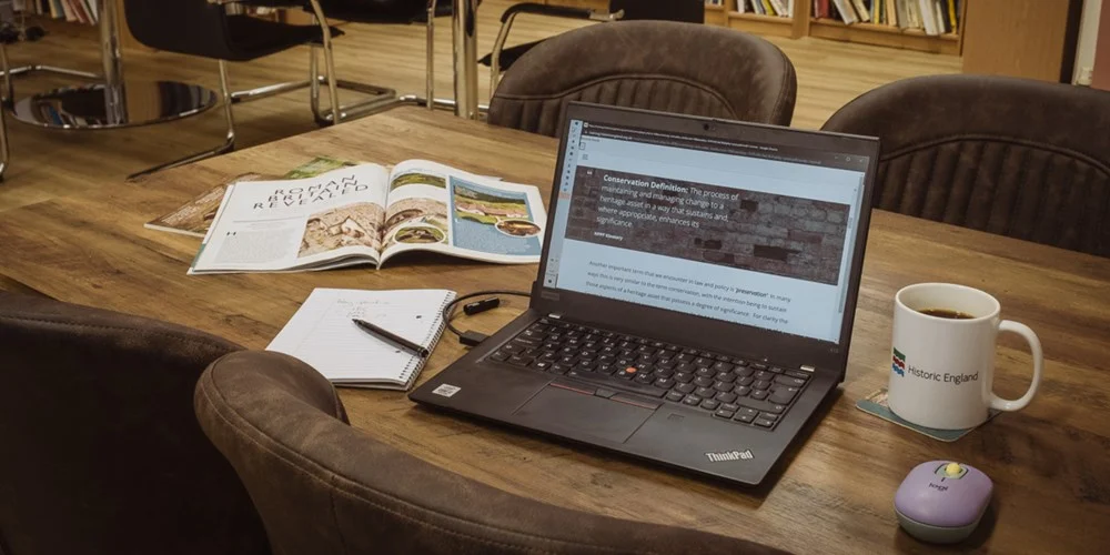 A laptop on a desk with a magazine, a notepad and pen, a mug, and a mouse