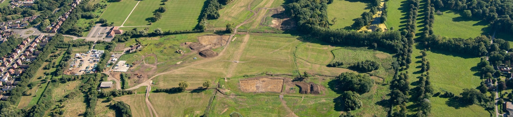 An aerial view of construction sites amid fields, with rows of houses in the background.