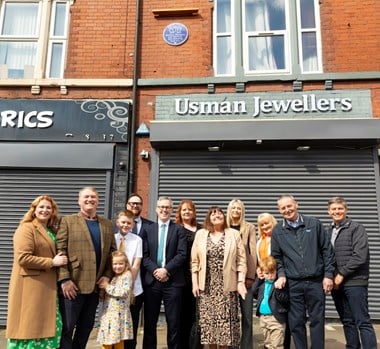 A family group of various ages stood in front of a red brick wall building with grey shutters and a blue heritage plaque installed next to a first storey window