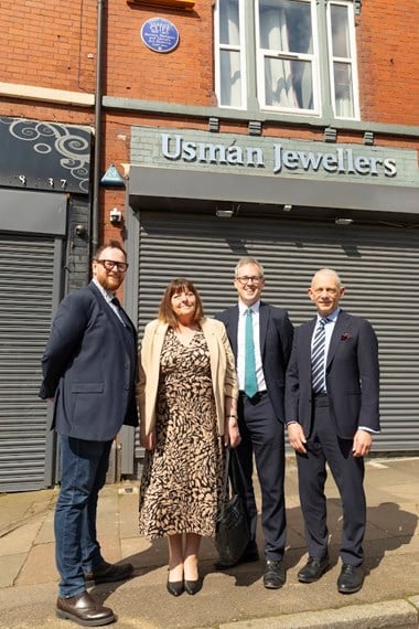 Four people stood in front of a red brick wall building with grey metal shutters and a blue plaque installed next to a first floor window