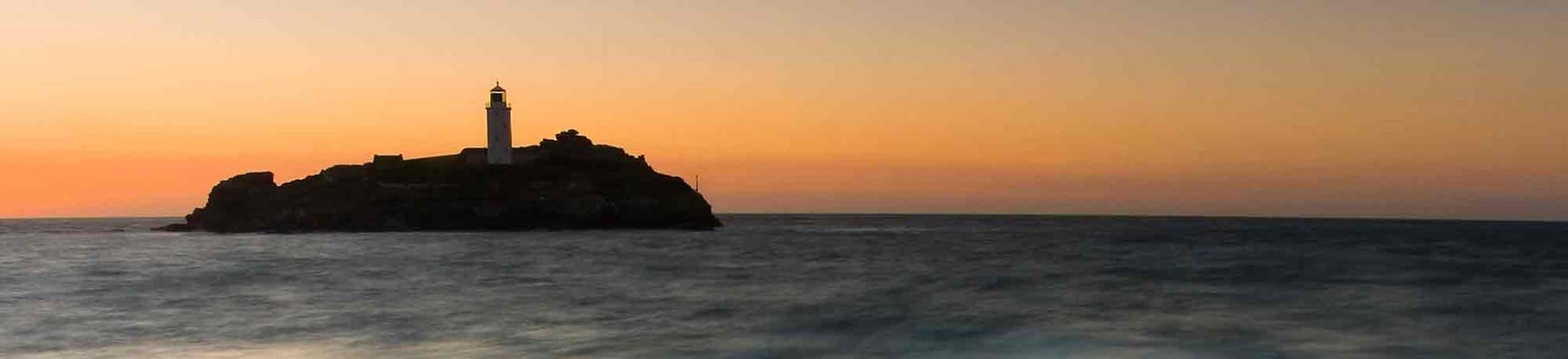 Lighthouse on the horizon with the sun setting behind. Rocks and white water in the foreground.