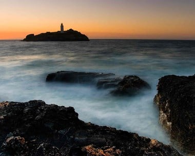 Lighthouse on the horizon with the sun setting behind. Rocks and white water in the foreground.