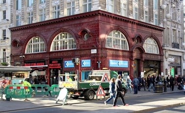 Red-tiled exterior of underground station with pedestrians in foreground on right and highway maintenance truck and works left.